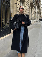 Langer schwarzer Mantel für Damen aus warmem Material, mit Taillengürtel und breiten Revers für einen eleganten Schnitt.