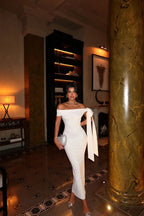 Robe longue blanche en tissu léger, épaules dénudées, coupe ajustée, avec un nœud décoratif pour une soirée élégante.
