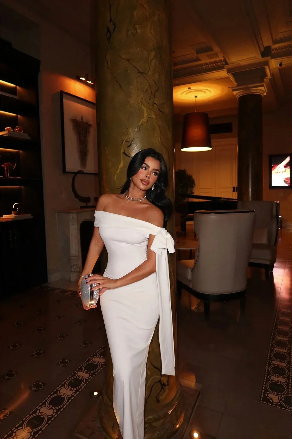 Robe longue blanche en tissu léger, épaules dénudées, coupe ajustée avec nœud détail pour une soirée élégante.