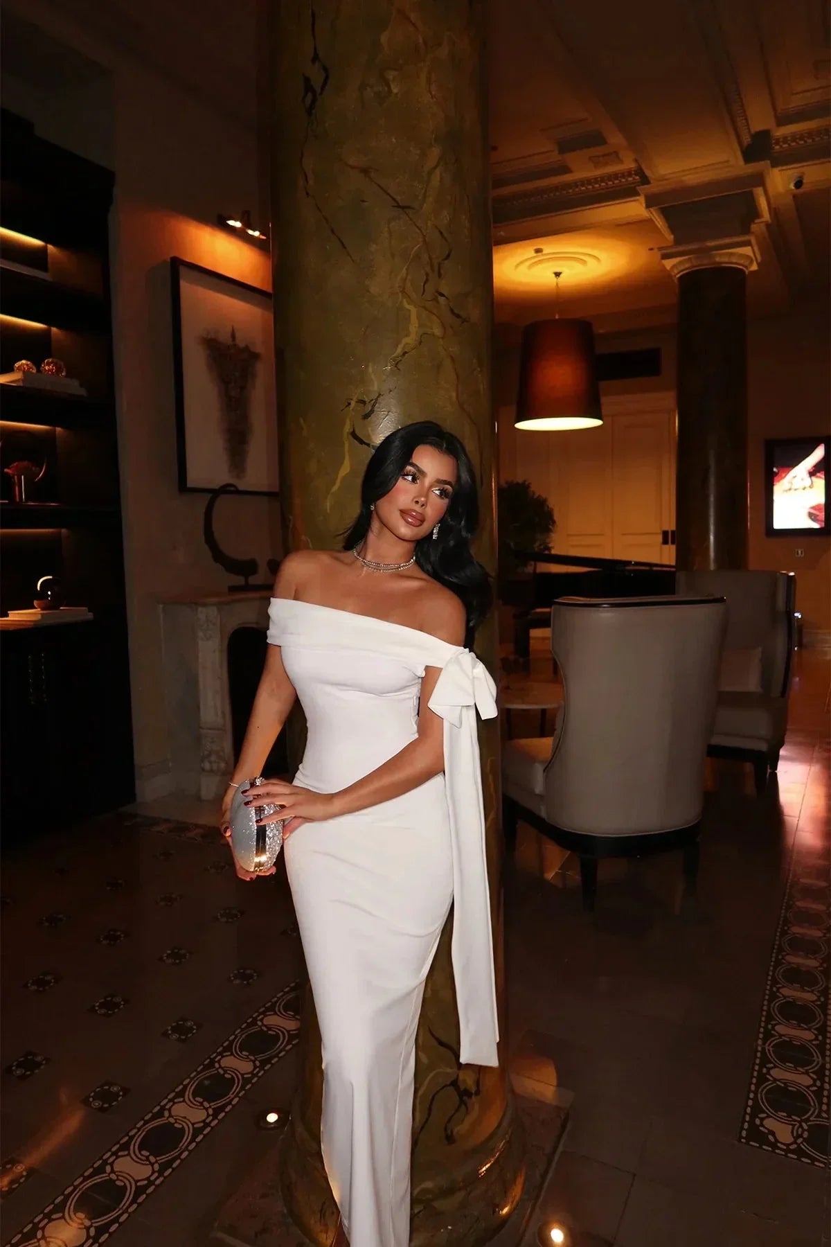 Robe longue blanche en tissu léger, épaules dénudées, coupe ajustée avec nœud détail pour une soirée élégante.